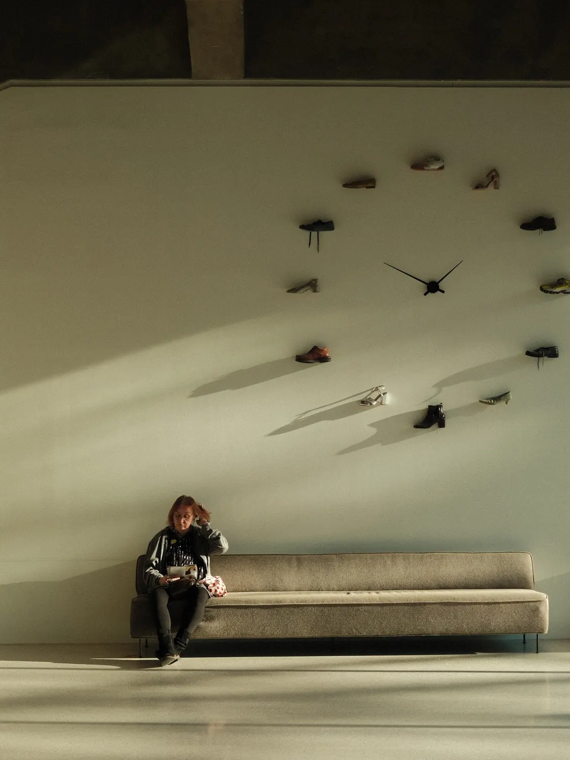 Woman seated alone on a long sofa beneath a wall of clocks
